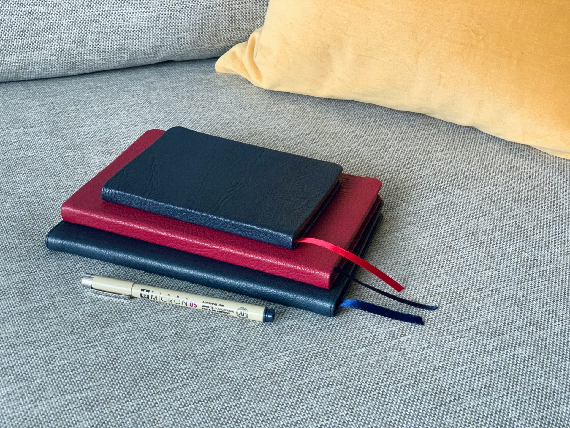 A stack of three closed notebooks with red and blue ribbon markers, and a pen, on a grey couch with a yellow cushion in the background.