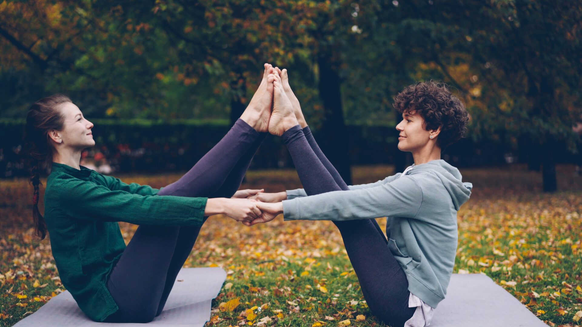 Two people practice partner yoga outdoors, sitting on mats in a park and balancing together with their feet touching and holding each other’s hands. Autumn leaves cover the grass around them.